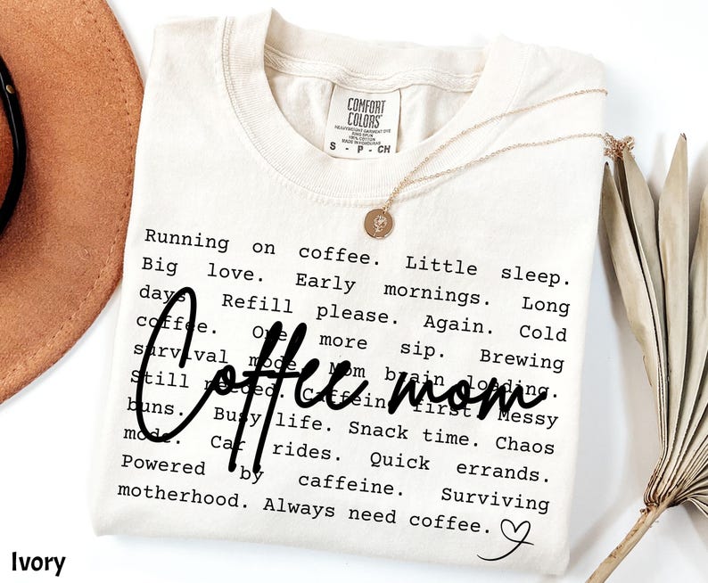 May include: Ivory-colored t-shirt with the words "Coffee mom" in a cursive font. The shirt has a list of coffee-related phrases in a black font. A gold necklace with a pendant is on the shirt.