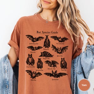 May include: A rust-colored t-shirt featuring a detailed "Bat Species Guide" graphic. The graphic includes illustrations of various bat species, such as the Little Brown Bat and the Free-tailed Bat. The shirt is made of a soft material and has a relaxed fit.