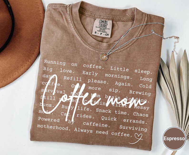 May include: Espresso-colored t-shirt with the words "Coffee mom" in a white script font. The shirt features a list of coffee-related phrases. A silver necklace with a small pendant rests on the shirt.