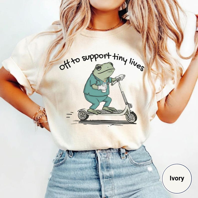 May include: Ivory-colored t-shirt with the text "off to support tiny lives." The graphic shows a frog in scrubs riding a scooter and holding a coffee cup. The shirt is paired with blue jeans.
