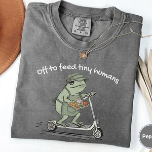 May include: A gray t-shirt with the phrase "off to feed tiny humans" and a cartoon frog riding a scooter, holding a tray of food. The shirt has a crew neck and short sleeves. A gold necklace is draped on the shirt.