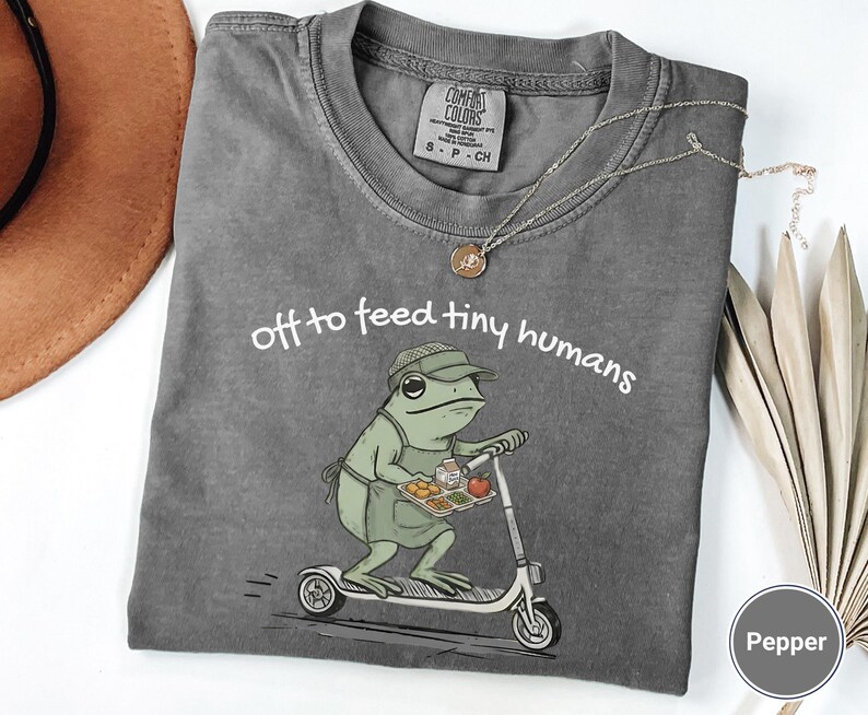 May include: A gray t-shirt with the phrase "off to feed tiny humans" and a cartoon frog riding a scooter, holding a tray of food. The shirt has a crew neck and short sleeves. A gold necklace is draped on the shirt.