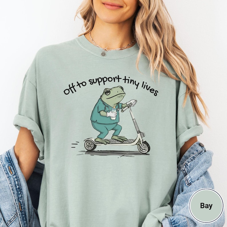 May include: A light green t-shirt with the phrase "off to support tiny lives" above a cartoon frog riding a scooter. The frog is wearing scrubs and holding a coffee cup. The scooter is white and gray.