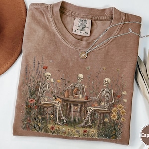 Może przedstawiać: Brązowy t-shirt z okrągłym dekoltem z nadrukiem graficznym przedstawiającym trzy szkielety siedzące przy stole na polu pełnym polnych kwiat&oacute;w, pijące kawę i jedzące ciasto. Koszulka posiada metkę marki Comfort Colors przy kołnierzu.