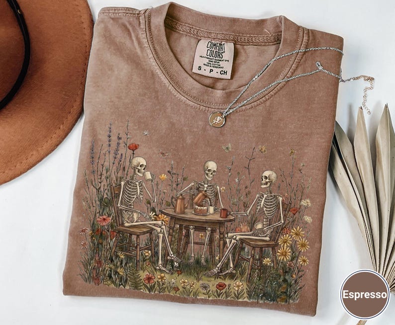 Może przedstawiać: Brązowy t-shirt z okrągłym dekoltem z nadrukiem graficznym przedstawiającym trzy szkielety siedzące przy stole na polu pełnym polnych kwiat&oacute;w, pijące kawę i jedzące ciasto. Koszulka posiada metkę marki Comfort Colors przy kołnierzu.