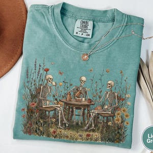 Może przedstawiać: Jasnozielony t-shirt z grafiką przedstawiającą trzy szkielety siedzące przy drewnianym stole na łące pełnej polnych kwiat&oacute;w, pijące kawę i jedzące ciasto. Koszulka posiada metkę marki Comfort Colors przy kołnierzu oraz oznaczenie rozmiaru S.