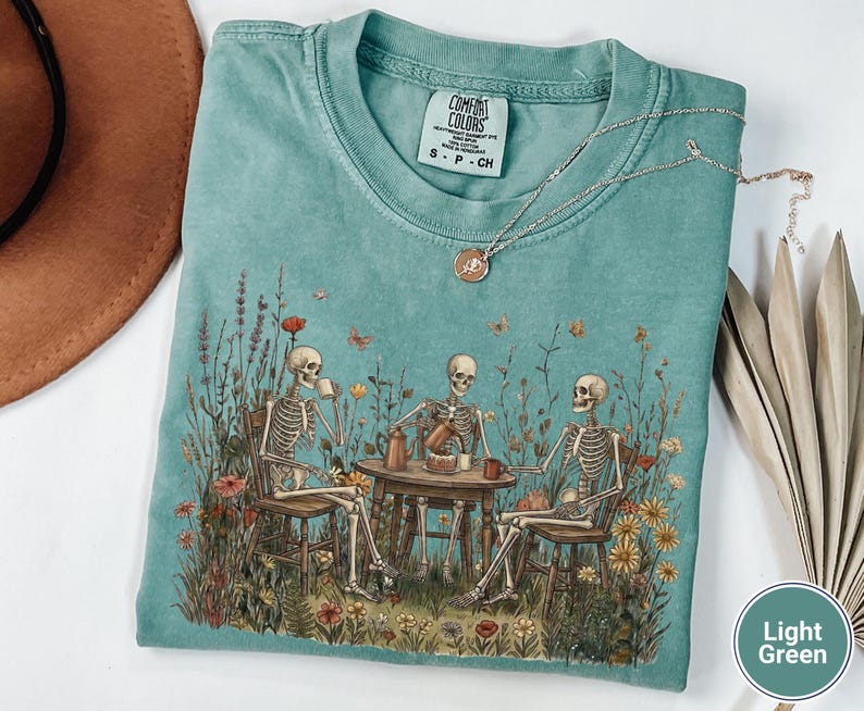 Może przedstawiać: Jasnozielony t-shirt z grafiką przedstawiającą trzy szkielety siedzące przy drewnianym stole na łące pełnej polnych kwiat&oacute;w, pijące kawę i jedzące ciasto. Koszulka posiada metkę marki Comfort Colors przy kołnierzu oraz oznaczenie rozmiaru S.