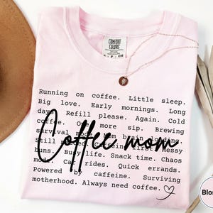 May include: A light pink t-shirt with the words "Coffee Mom" in a handwritten font. The shirt has a list of phrases about motherhood and coffee. A gold necklace with a pendant is on the shirt.