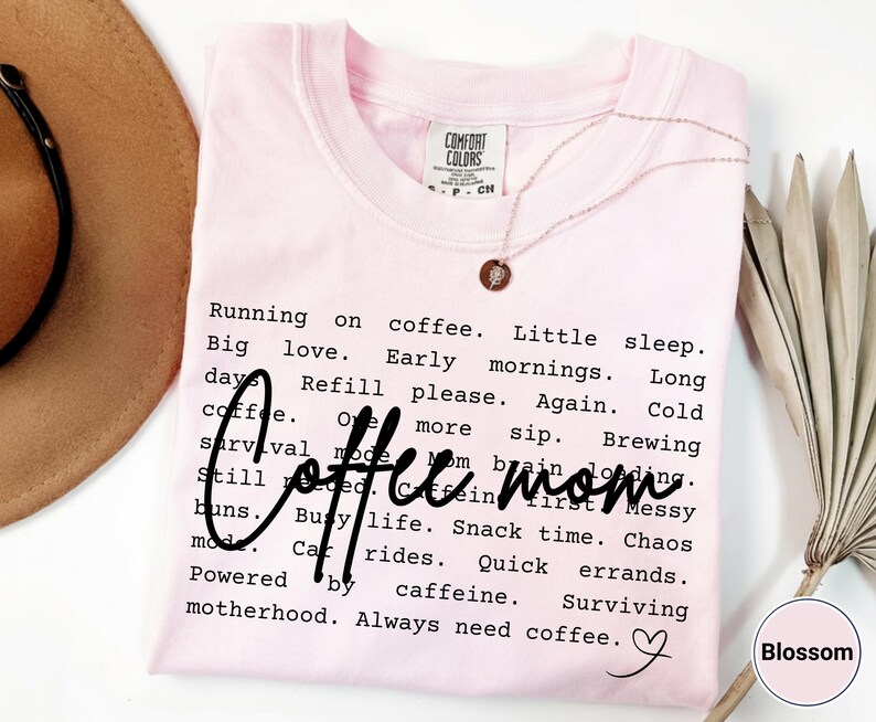 May include: A light pink t-shirt with the words "Coffee Mom" in a handwritten font. The shirt has a list of phrases about motherhood and coffee. A gold necklace with a pendant is on the shirt.