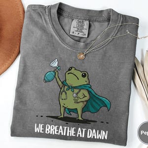 Pode incluir: Uma t-shirt cinza-carv&atilde;o com o gr&aacute;fico de um sapo verde vestindo uma capa azul-petr&oacute;leo e um estetosc&oacute;pio, segurando um ressuscitador manual azul. O texto abaixo do sapo diz WE BREATHE AT DAWN. Etiqueta da marca Comfort Colors.