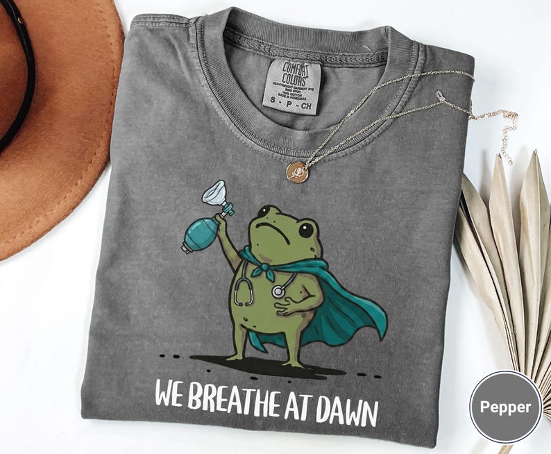 Pode incluir: Uma t-shirt cinza-carv&atilde;o com o gr&aacute;fico de um sapo verde vestindo uma capa azul-petr&oacute;leo e um estetosc&oacute;pio, segurando um ressuscitador manual azul. O texto abaixo do sapo diz WE BREATHE AT DAWN. Etiqueta da marca Comfort Colors.