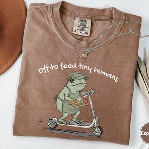 May include: Espresso-colored Comfort Colors t-shirt featuring a frog riding a scooter, carrying a tray of food. The text "off to feed tiny humans" is printed above the frog. A silver necklace with a pendant is draped across the shirt.