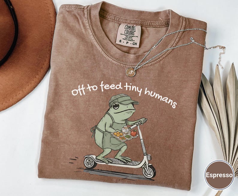 May include: Espresso-colored Comfort Colors t-shirt featuring a frog riding a scooter, carrying a tray of food. The text "off to feed tiny humans" is printed above the frog. A silver necklace with a pendant is draped across the shirt.