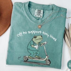 May include: Light green t-shirt with the text "off to support tiny lives" and a cartoon frog riding a scooter. The frog is wearing scrubs and holding a baby bottle. A silver necklace with a pendant is on the shirt.
