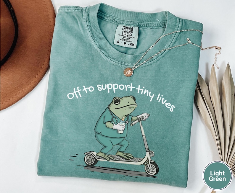 May include: Light green t-shirt with the text "off to support tiny lives" and a cartoon frog riding a scooter. The frog is wearing scrubs and holding a baby bottle. A silver necklace with a pendant is on the shirt.