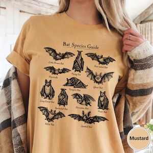 May include: Mustard-colored t-shirt featuring a black and white illustration of various bat species. The design includes the text "Bat Species Guide" and names like "Little Brown Bat" and "Flying Fox". The shirt is a casual wear item.