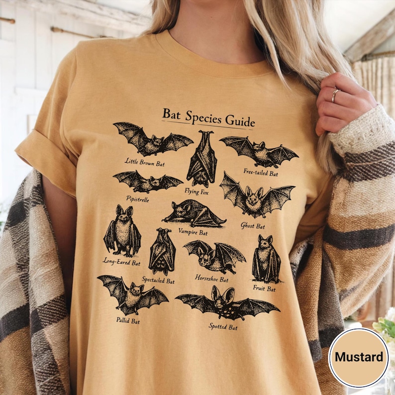 May include: Mustard-colored t-shirt featuring a black and white illustration of various bat species. The design includes the text "Bat Species Guide" and names like "Little Brown Bat" and "Flying Fox". The shirt is a casual wear item.