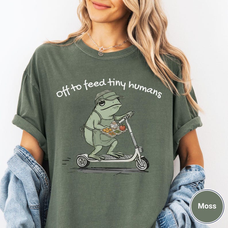 May include: Moss green t-shirt with the phrase "off to feed tiny humans" in white. The graphic features a frog riding a scooter while holding a tray of food. The frog wears a hat and apron.