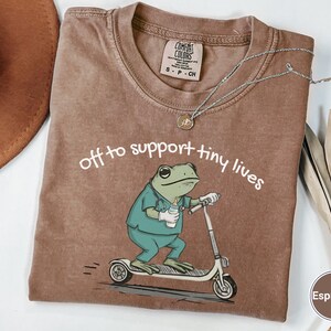 May include: Espresso-colored t-shirt with the phrase "off to support tiny lives" and a cartoon frog riding a scooter. The frog is wearing scrubs and holding a bottle. A silver necklace and a brown hat are also visible.