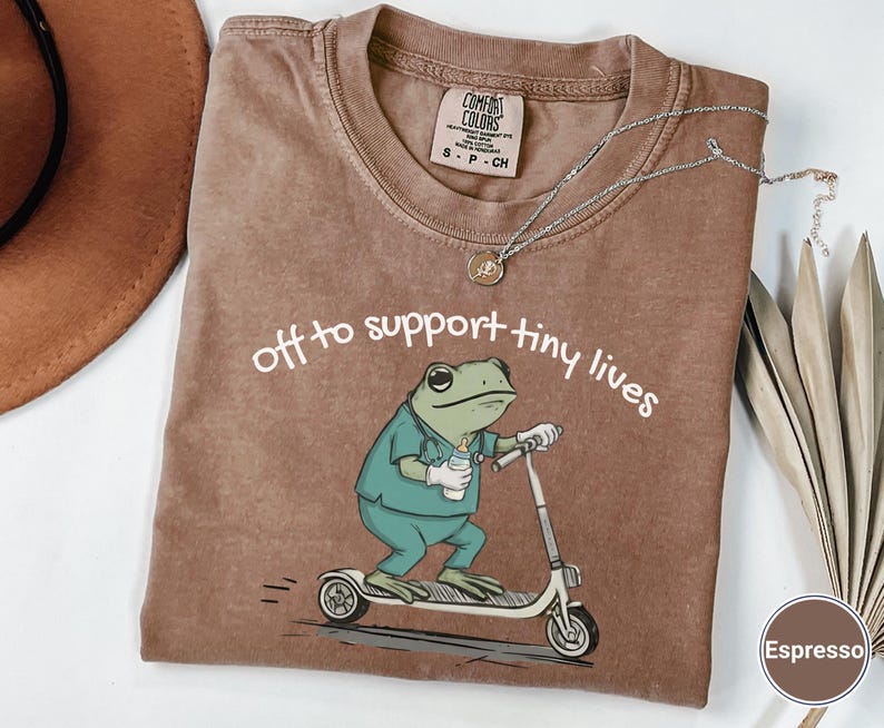 May include: Espresso-colored t-shirt with the phrase "off to support tiny lives" and a cartoon frog riding a scooter. The frog is wearing scrubs and holding a bottle. A silver necklace and a brown hat are also visible.
