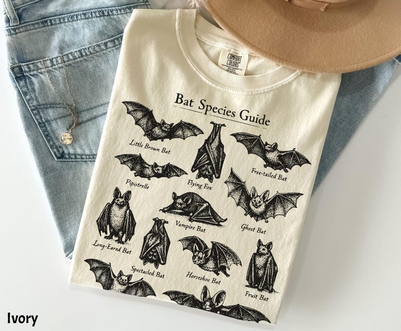 May include: Ivory-colored t-shirt featuring a "Bat Species Guide" graphic with various bat illustrations and names. The shirt is laid on top of blue jeans and a tan hat. The text "Ivory" is at the bottom left.
