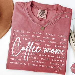 May include: Crimson-colored t-shirt with the words "Coffee mom" in a white script font. The shirt features a list of coffee-related phrases and a small heart graphic. A silver necklace with a pendant is draped on the shirt.