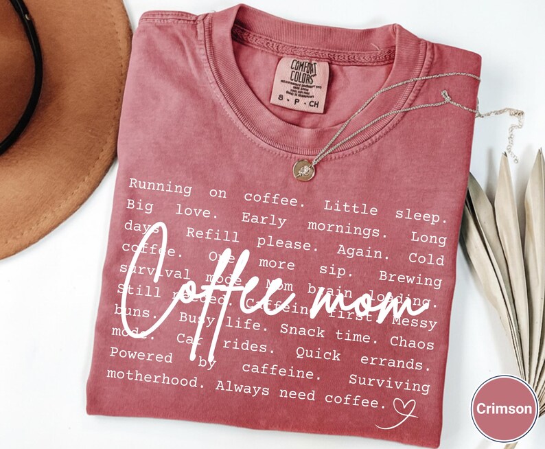 May include: Crimson-colored t-shirt with the words "Coffee mom" in a white script font. The shirt features a list of coffee-related phrases and a small heart graphic. A silver necklace with a pendant is draped on the shirt.