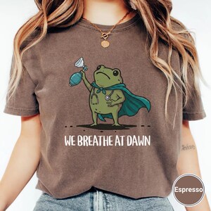 Pode incluir: Uma t-shirt castanha com um gr&aacute;fico de um sapo verde a usar uma capa azul-esverdeada e um estetosc&oacute;pio, segurando um bal&atilde;o de reanima&ccedil;&atilde;o. Abaixo do sapo, o texto diz WE BREATHE AT DAWN. Uma etiqueta circular exibe a palavra Espresso.