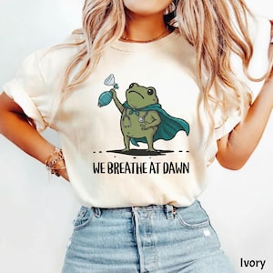Pode incluir: Uma t-shirt cor de marfim com um gr&aacute;fico de um sapo verde a usar uma capa azul-petr&oacute;leo e um estetosc&oacute;pio, segurando um bal&atilde;o de reanima&ccedil;&atilde;o. Abaixo do sapo, o texto WE BREATHE AT DAWN est&aacute; escrito em letras mai&uacute;sculas pretas.