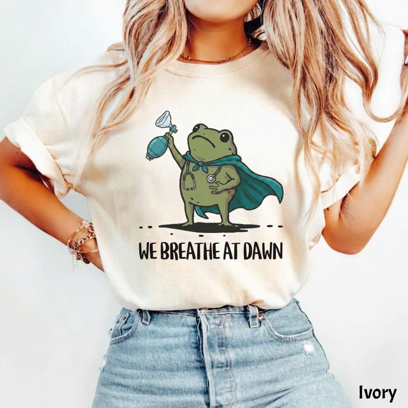 Pode incluir: Uma t-shirt cor de marfim com um gr&aacute;fico de um sapo verde a usar uma capa azul-petr&oacute;leo e um estetosc&oacute;pio, segurando um bal&atilde;o de reanima&ccedil;&atilde;o. Abaixo do sapo, o texto WE BREATHE AT DAWN est&aacute; escrito em letras mai&uacute;sculas pretas.