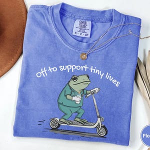 May include: Flo Blue t-shirt with the text "off to support tiny lives" and a cartoon frog riding a scooter. The frog is wearing scrubs and holding a baby bottle. A gold necklace and a brown hat are also visible.