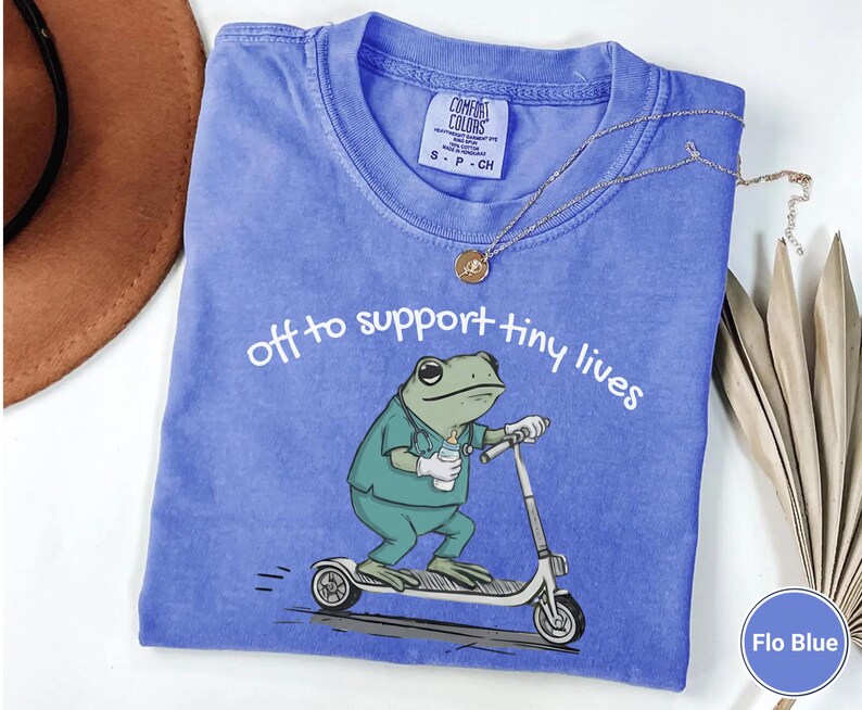 May include: Flo Blue t-shirt with the text "off to support tiny lives" and a cartoon frog riding a scooter. The frog is wearing scrubs and holding a baby bottle. A gold necklace and a brown hat are also visible.
