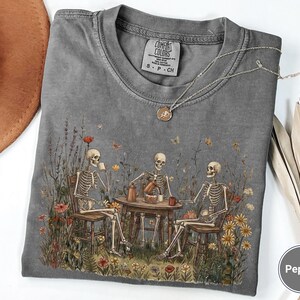 Może przedstawiać: Ciemnoszary t-shirt z okrągłym dekoltem z nadrukiem trzech szkielet&oacute;w siedzących przy drewnianym stole na polu dzikich kwiat&oacute;w i pijących z kubk&oacute;w. Koszulka posiada metkę Comfort Colors oraz okrągłą etykietę z napisem Pepper.
