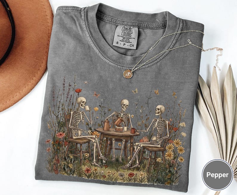 Może przedstawiać: Ciemnoszary t-shirt z okrągłym dekoltem z nadrukiem trzech szkielet&oacute;w siedzących przy drewnianym stole na polu dzikich kwiat&oacute;w i pijących z kubk&oacute;w. Koszulka posiada metkę Comfort Colors oraz okrągłą etykietę z napisem Pepper.