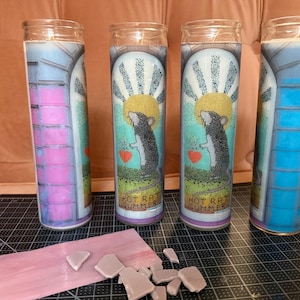 May include: Four glass candles with colorful designs. Two candles feature a rat illustration with a heart, sun, and text "HOT RAT SUMMER." The other two candles have a stained glass design with pink and blue hues. A pink object with broken pieces is in the foreground.