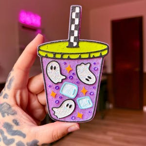 May include: A purple cup-shaped decoration featuring a lime green lid and a black and white checkered straw. The cup contains white glittery ghost shapes, light blue ice cubes, small purple beads, and orange star accents on a textured surface.
