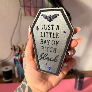 May include: A coffin-shaped decoration with the text "JUST A LITTLE RAY OF PITCH Black". The decoration is silver with black glitter accents, a bat, and blue gem embellishments. The edges are outlined in black.