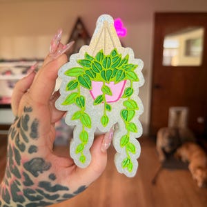 May include: A decorative plant-shaped item with a glittery, scalloped edge. The design features a pink pot with green leaves and vines. The plant is suspended from a gold-coloured hanger. The item is held in a hand with a leopard print tattoo.