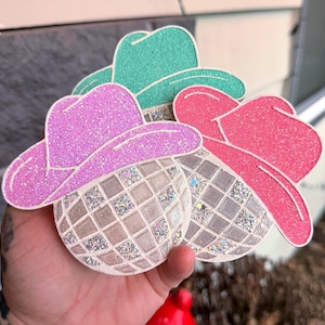 May include: Three glittery cowboy hat ornaments in pink, green, and red. Each hat sits atop a disco ball-shaped base with a grid pattern. The ornaments are held in a hand, suggesting a craft or party theme.