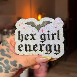 May include: A white glittery sticker with a scalloped edge featuring a black bat icon and the text hex girl energy in a black gothic font. Small pink star accents are scattered around the bat, creating a decorative and textured appearance.