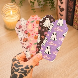 May include: Four rectangular bookmarks with scalloped tops featuring Halloween patterns. One is light pink with ghosts and pumpkins, one is pink with candy corn, one is black with ghosts, and one is purple with white ghosts and the text Boo.