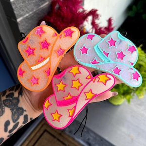 May include: Three cowboy hat-shaped ornaments in orange, light blue, and pink. Each hat features a star pattern in contrasting colors: pink stars on the orange hat, pink stars on the light blue hat, and yellow stars on the pink hat. Black string attached.