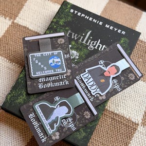 May include: Three rectangular magnetic bookmarks featuring Twilight-themed designs. One shows a sign reading The City of Forks Welcomes You, another depicts a character with the text Daddy, and the third features a silhouette with the quote This is the skin of a killer, Bella. The bookmarks are displayed on a copy of the book Twilight by Stephenie Meyer.