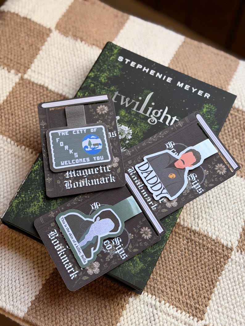 May include: Three rectangular magnetic bookmarks featuring Twilight-themed designs. One shows a sign reading The City of Forks Welcomes You, another depicts a character with the text Daddy, and the third features a silhouette with the quote This is the skin of a killer, Bella. The bookmarks are displayed on a copy of the book Twilight by Stephenie Meyer.