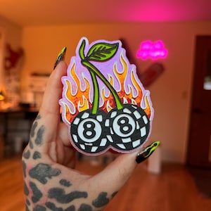 May include: A sticker featuring two cherries with the number 8 on each, designed to look like billiard balls. The cherries are surrounded by flames and have a green stem and leaf. The background is a light purple.