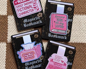 May include: Four rectangular magnetic bookmarks featuring black covers with floral patterns and white text. Each bookmark displays a unique pink graphic: a warning sign, a book, a tissue box labeled Crying Over My Book Boyfriend, and a typewriter.