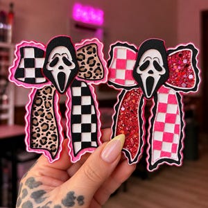 May include: Two decorative bows featuring the Ghostface mask from the Scream franchise. The left bow has black and white checkered patterns and leopard print. The right bow features pink and white checkered patterns and red glitter accents.