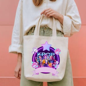 May include: A cream-coloured tote bag with a colourful graphic. The design features the text "KPOP DESIGN", a purple star, musical notes, and images of three women. The bag is held by a person.
