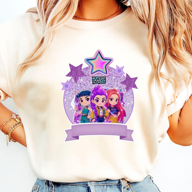 May include: A cream-coloured t-shirt with a graphic of three cartoon characters with purple, pink, and blue hair. The design includes a purple glittery background, stars, and the text "KPOP DEMON HUNTERS". A purple banner is at the bottom.