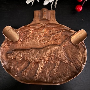 May include: A bronze-colored ashtray with a detailed relief of a dog in a field. The ashtray has two built-in rests and a rectangular top. The overall design is oval-shaped.
